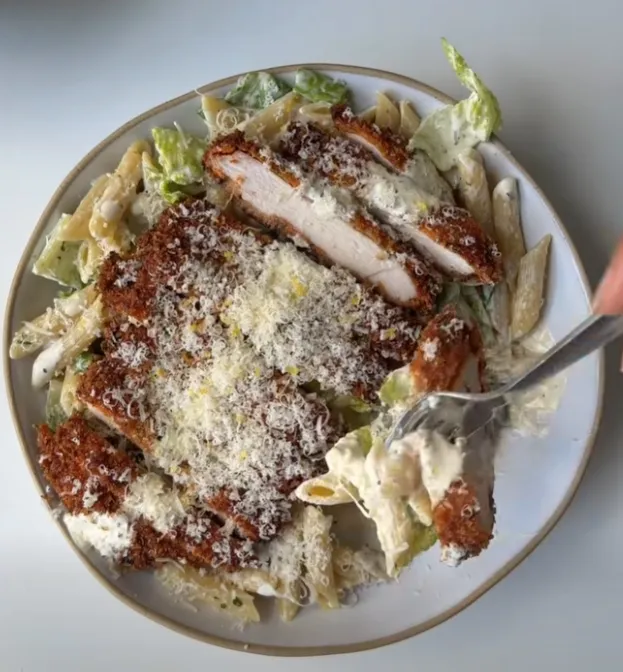 Crispy breaded chicken sliced over caesar penne salad with a fork lifting some pasta