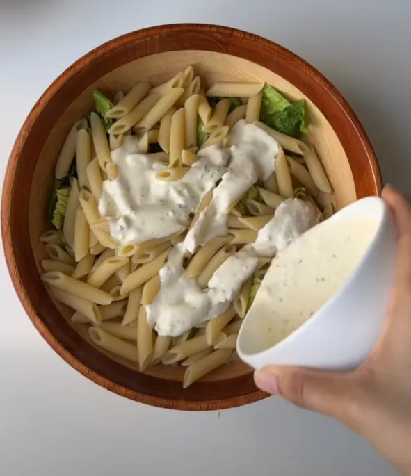 Caesar dressing being poured over penne and romaine in a wooden bowl
