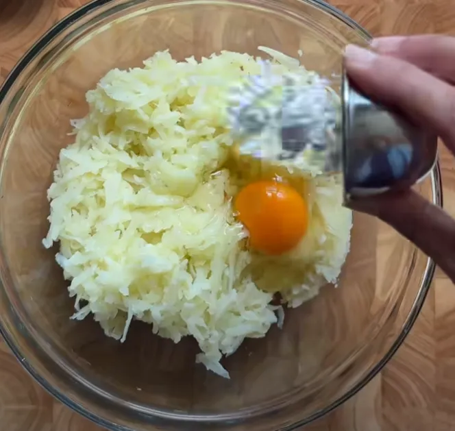 An egg being added to grated potato in a glass bowl
