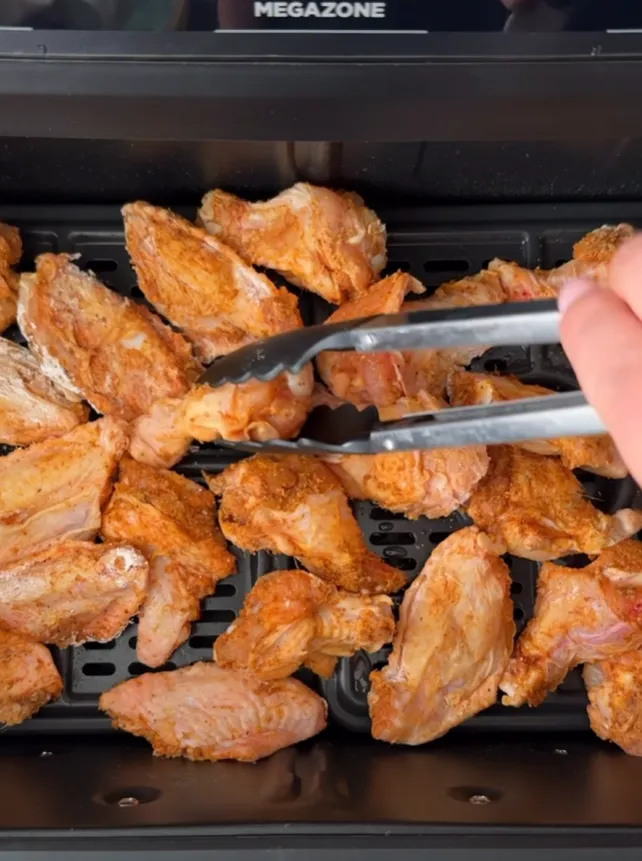 Seasoned chicken wings arranged in an air fryer basket being turned with tongs