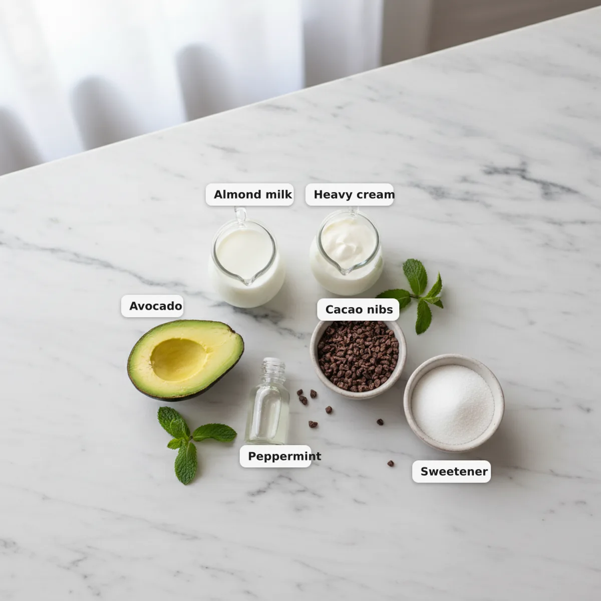 All the ingredients for a keto mint chocolate chip smoothie laid out and labelled on a marble surface — almond milk, heavy cream, avocado, peppermint extract, cacao nibs and sweetener
