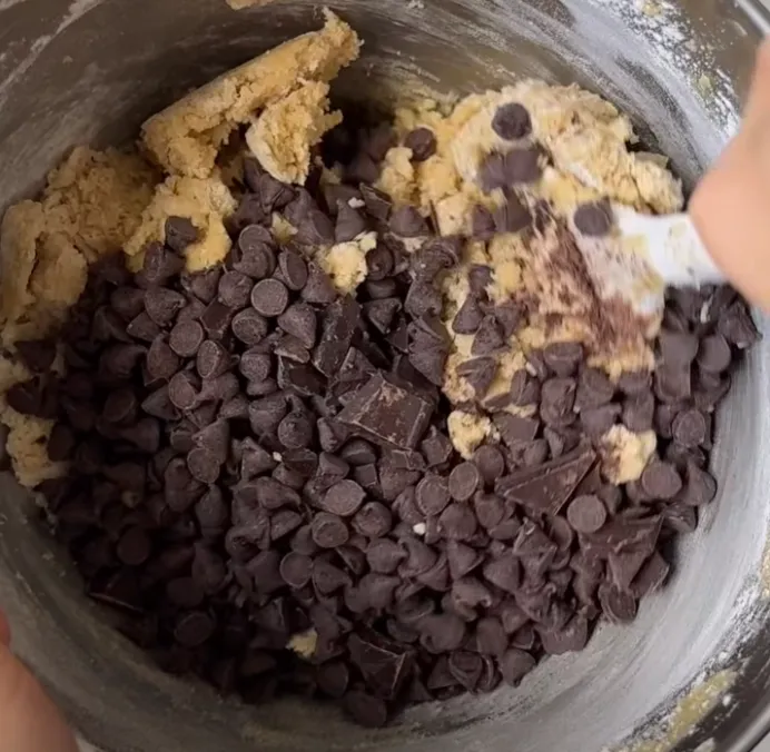 Chocolate chips and chopped chocolate being folded into thick cookie dough in a bowl