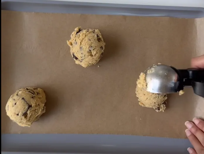 Large balls of chocolate chip cookie dough being scooped onto a lined tray
